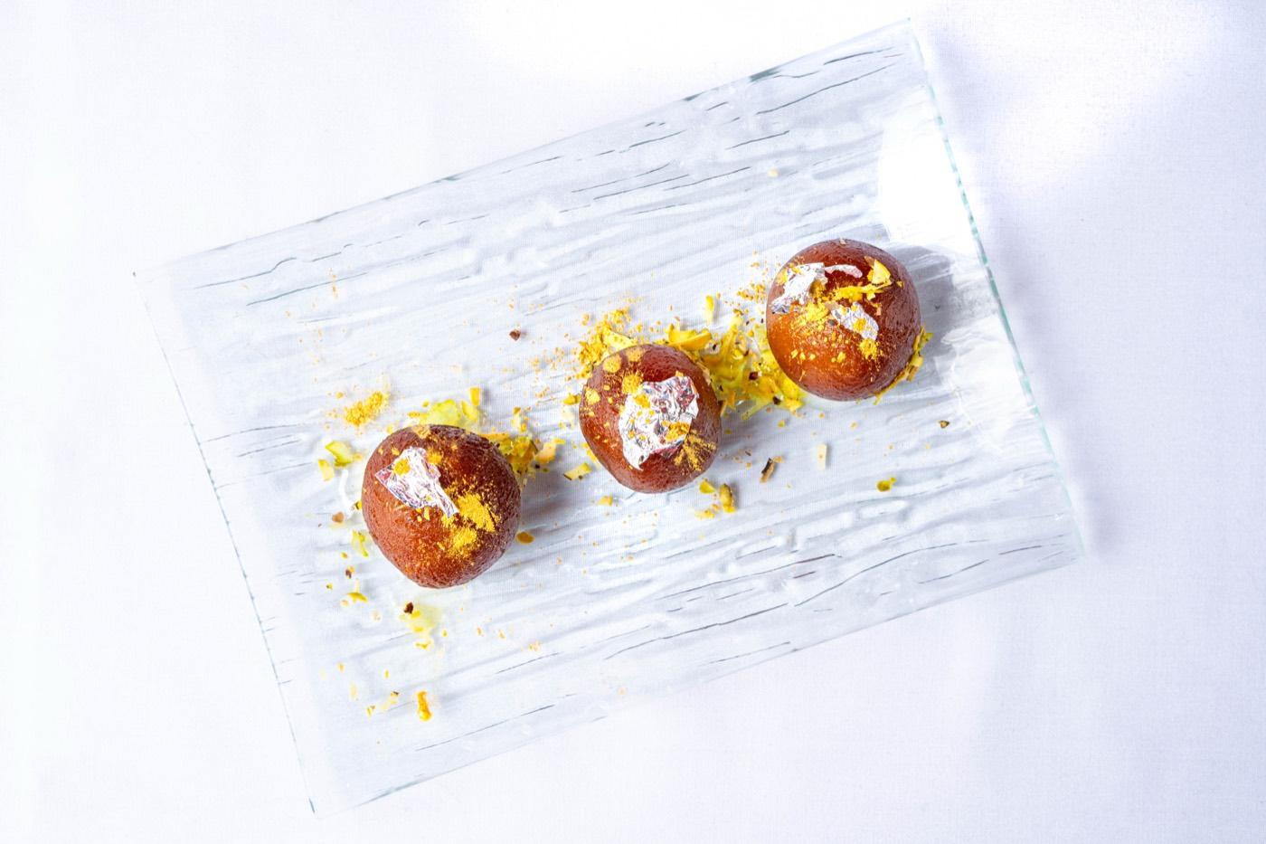 Three gulab jamun topped with silver leaf on a glass plate at India 101 in Irving, TX